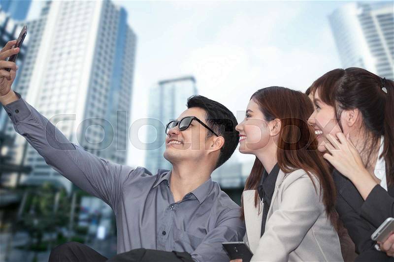 Businessman sitting and using phone to take photo as selfie with colleague together and modern office building background, stock photo