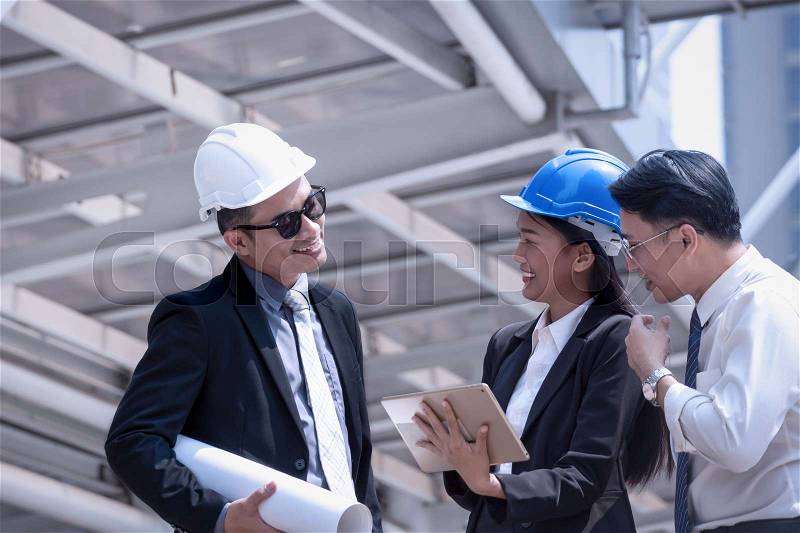 Asian businessman and woman discuss with engineer architect professional wearing safety helmet and suite, looking at tablet and holding construction roll plan for working team concept, stock photo