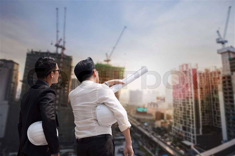 Asian businessman discuss with engineer architect professional wearing safety helmet and suite, looking and point to finger forward away and holding construction roll plan for working team concept, stock photo