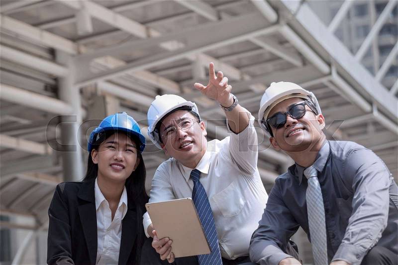 Asian businessman and woman discuss with engineer architect professional wearing safety helmet and suite, looking follow point to finger and tablet in hand for working team concept, stock photo