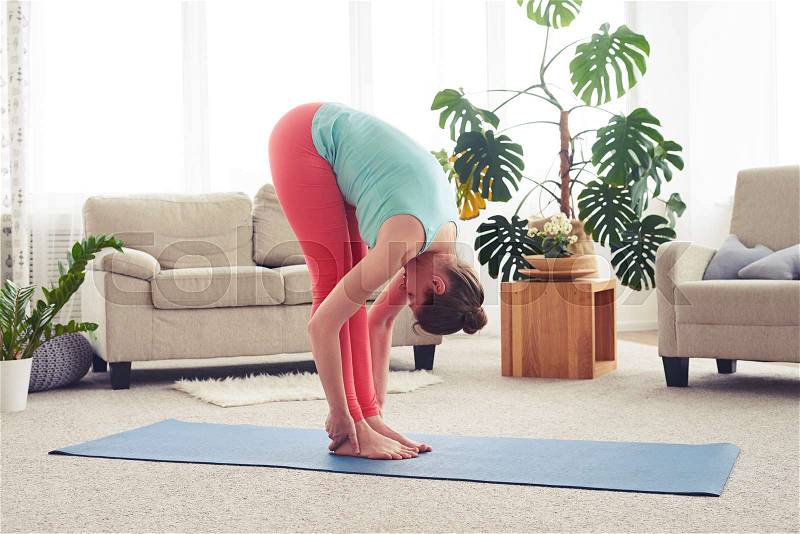 Wide shot of young woman stretching on yoga mate in living room, stock photo