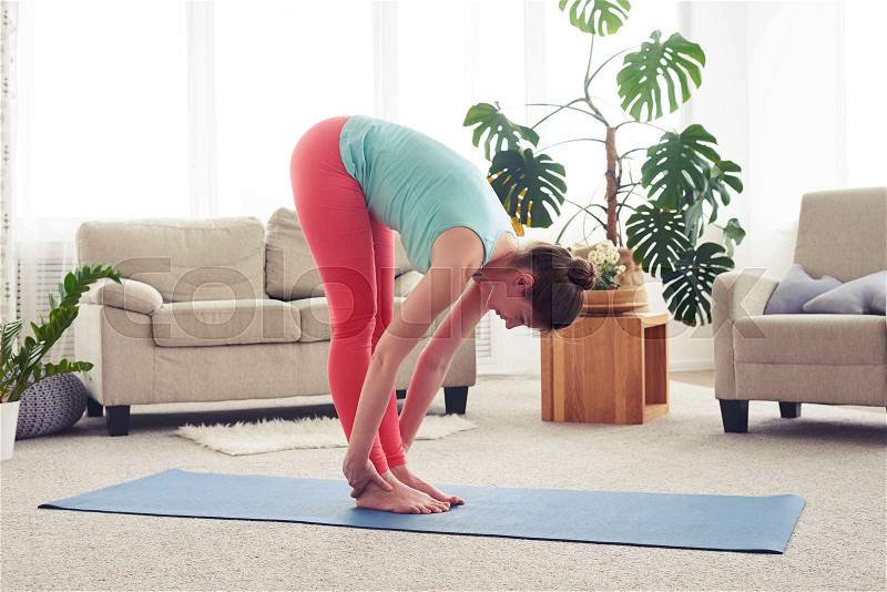 Wide shot of skinny female stretching on yoga mate in living room, stock photo