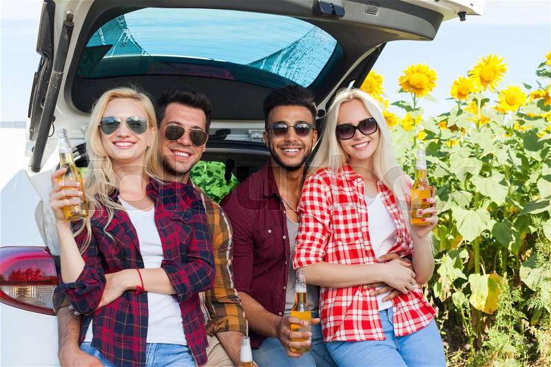 Friends drinking beer toasting clink bottles sitting in car trunk outdoor countryside, happy smile people group summer sunflower field, stock photo