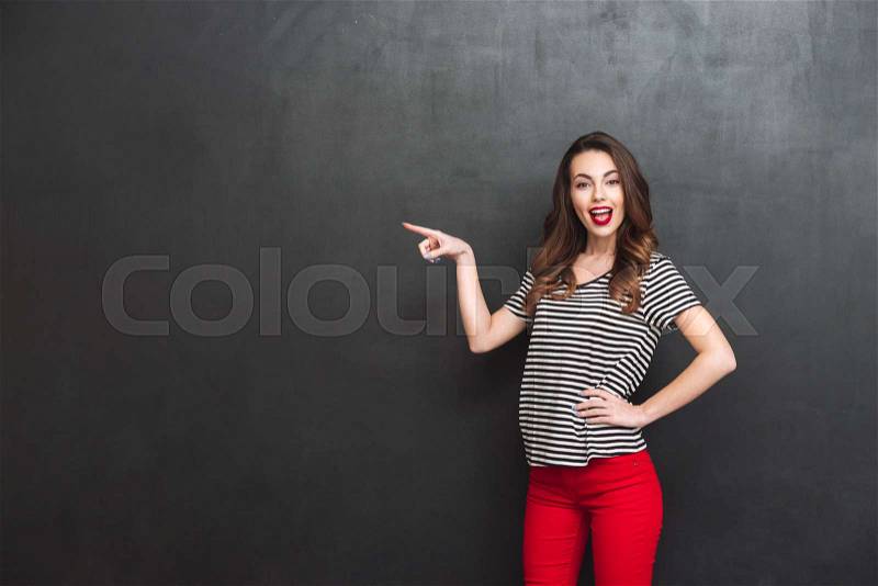 Happy woman pointing away and holding arm at hip while looking at the camera over black background, stock photo