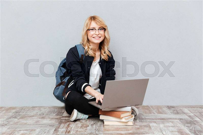 Image of happy young woman student wearing glasses with backpack posing over grey wall. Looking at camera using laptop computer, stock photo
