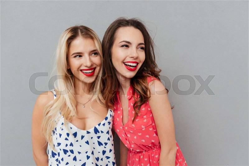 Picture of happy young two ladies friends with bright makeup lips standing over grey wall and posing. Looking aside, stock photo