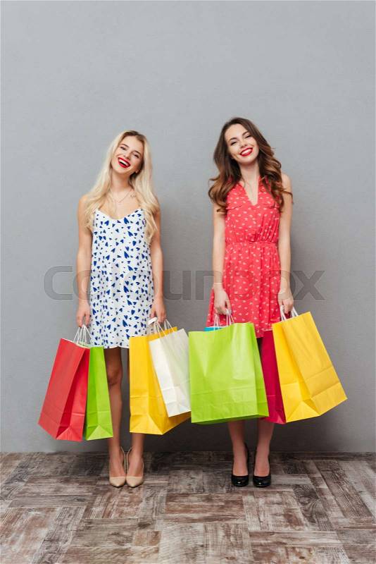 Image of cheerful young two ladies friends with bright makeup lips standing over grey wall and posing with shopping bags. Looking at camera, stock photo