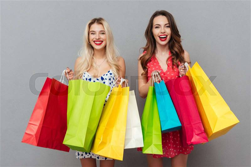 Picture of happy young two ladies friends with bright makeup lips standing over grey wall and posing with shopping bags. Looking at camera, stock photo