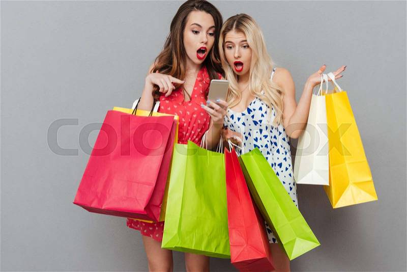 Photo of surprised young two ladies friends with bright makeup lips standing over grey wall and posing with shopping bags using mobile phone. Looking aside, stock photo