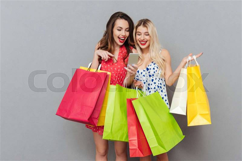 Image of happy young two ladies friends with bright makeup lips standing over grey wall and posing with shopping bags using mobile phone. Looking aside, stock photo