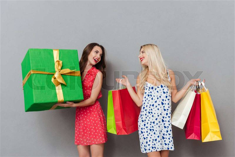Photo of amazing young two ladies friends with bright makeup lips standing over grey wall and posing with shopping bags and gift. Looking aside, stock photo