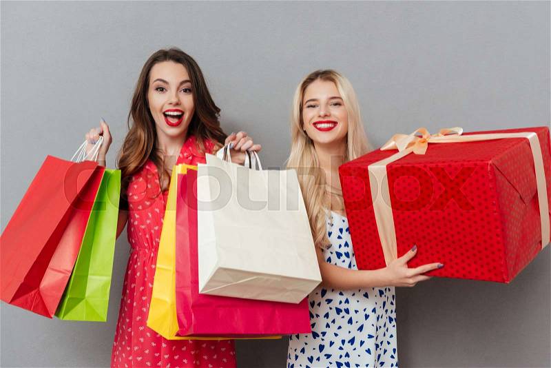 Image of cheerful young two ladies friends with bright makeup lips standing over grey wall and posing with shopping bags and gift. Looking at camera, stock photo