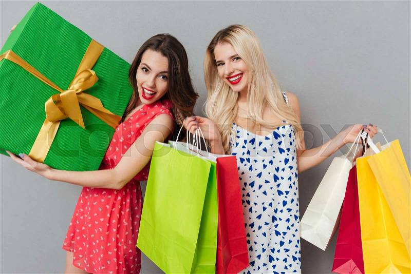 Photo of smiling young two ladies friends with bright makeup lips standing over grey wall and posing with shopping bags and gift. Looking at camera, stock photo