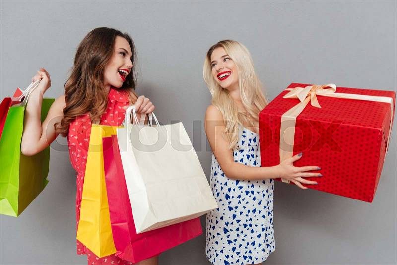 Image of amazing young two ladies friends with bright makeup lips standing over grey wall and posing with shopping bags and gift. Looking aside, stock photo