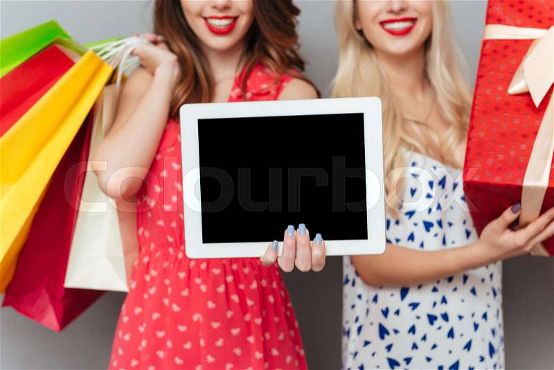 Cropped image of happy young two ladies friends with bright makeup lips standing over grey wall and posing with shopping bags and gift. Showing display of tablet computer, stock photo