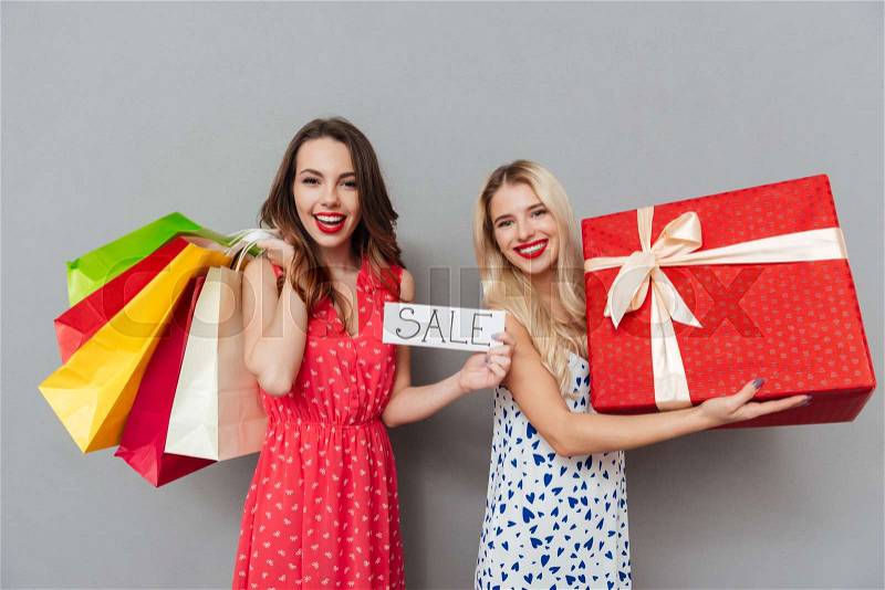 Photo of happy young two ladies friends with bright makeup lips standing over grey wall and posing with shopping bags and gift. Looking at camera, stock photo