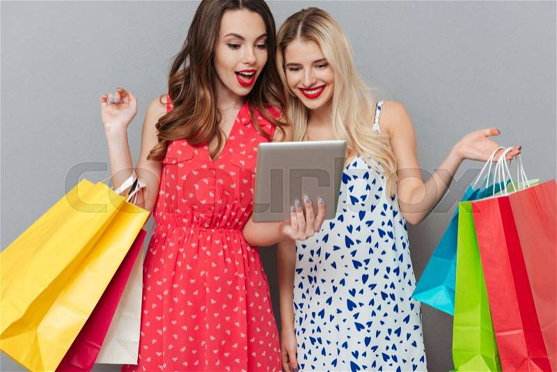 Image of cheerful emotional young two ladies friends with bright makeup lips standing over grey wall and posing with shopping bags. Looking at tablet computer, stock photo