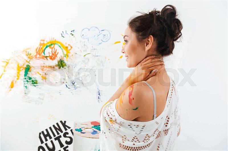 Smiling attractive young woman with brushes on palette standing and painting over white background, stock photo