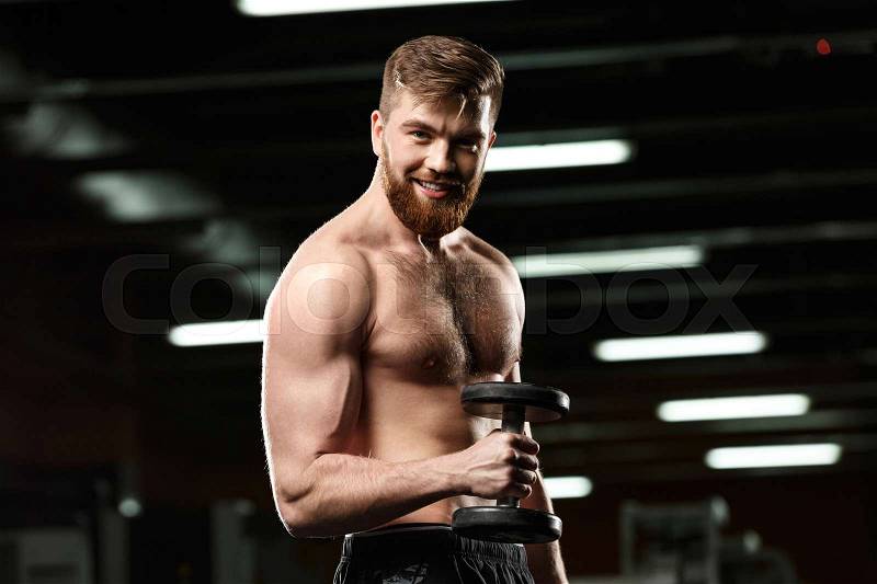 Image of happy sports man make sport exercises with dumbbells in gym and looking at camera, stock photo