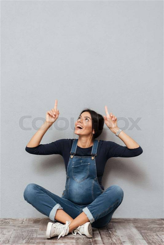 Image of happy pregnant lady sitting on floor over grey background pointing to copyspace. Looking aside, stock photo