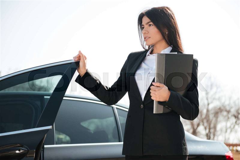 Beautiful young businesswoman holding folder standing near the car, stock photo