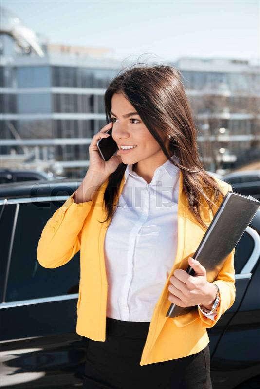 Happy young businesswoman in yellow jacket holding folder and talking on mobile phone outdoors, stock photo