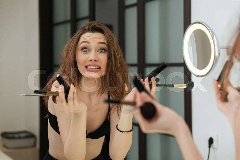 Confused pretty young woman holding brushes and doing makeup in bathroom, stock photo