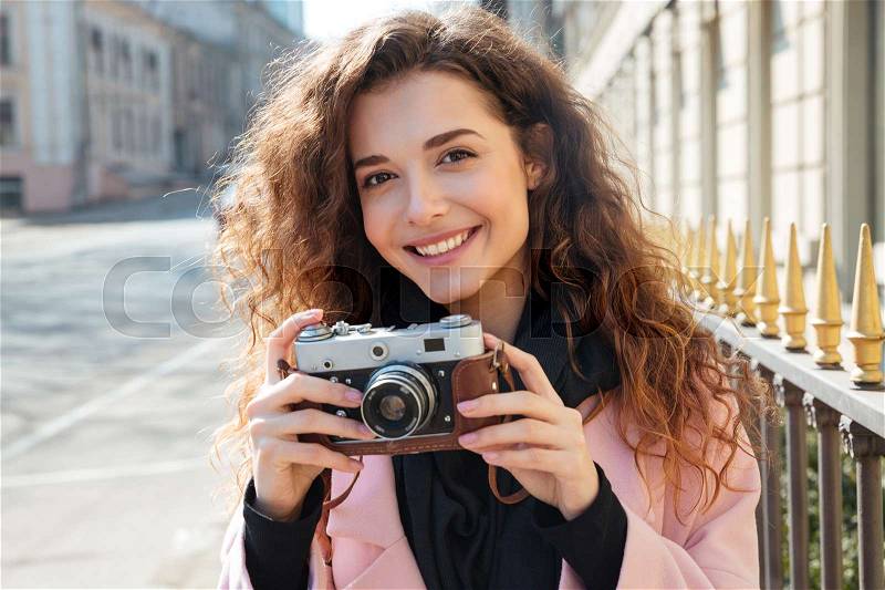 Picture of a young woman in coat which holding retro camera in hands and looking at the camera, stock photo