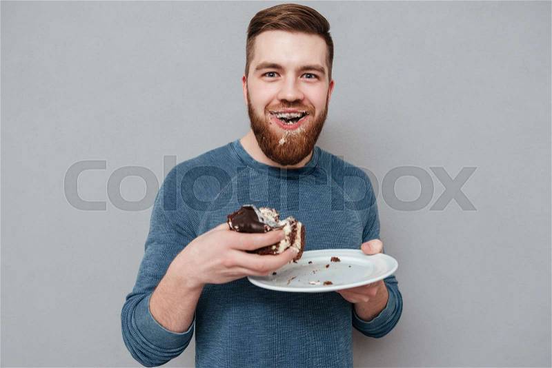 Filthy bearded young man eating chocolate cakes isolated on gray background, stock photo