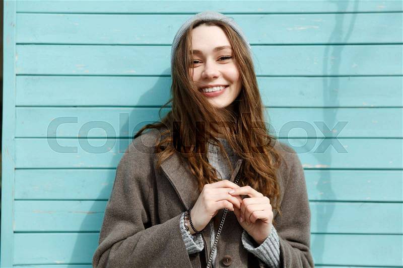 Portrait of happy beautiful young woman in hat and coat standing over blue wall background, stock photo