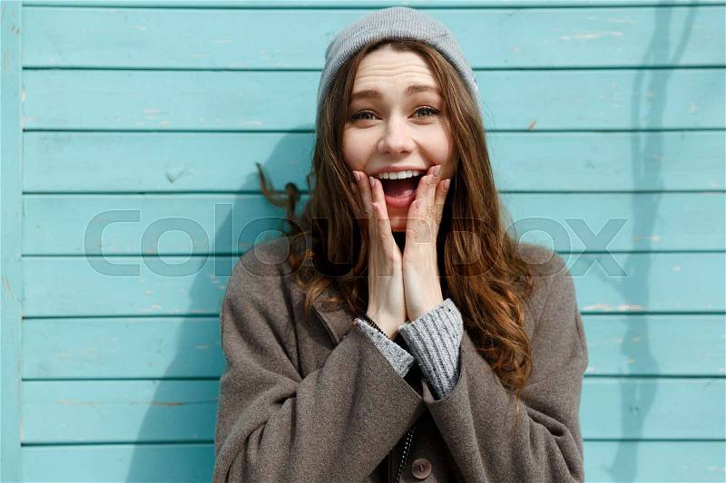 Surprised cheerful young woman in hat standing and shouting over blue wall background, stock photo