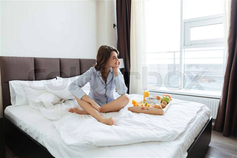 Attractive young woman having breakfast sitting on bed in hotel and looking away, stock photo