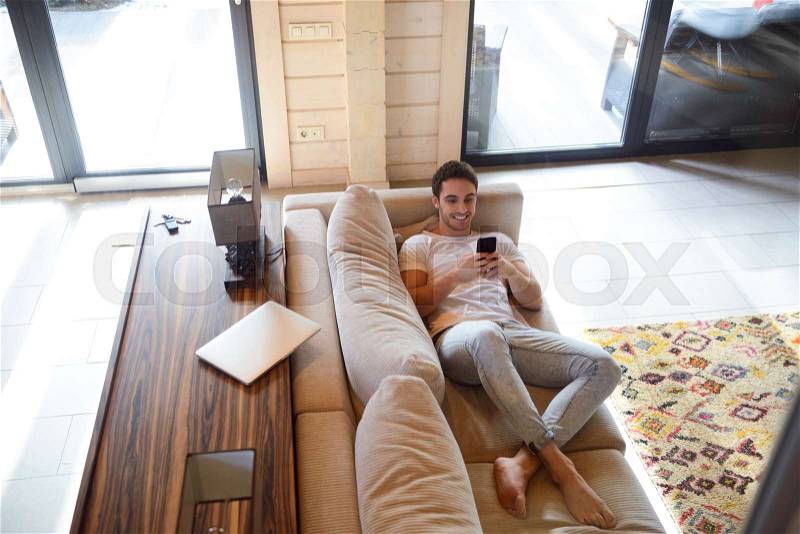 Full-length shot of cheerful man with phone lying on soft couch, stock photo