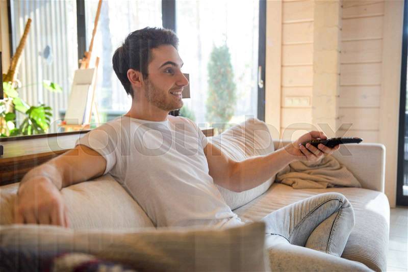 Sideview of sitting on couch man with remote controller in hand, stock photo