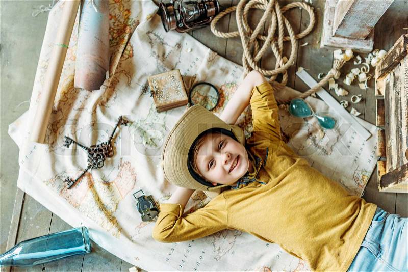 Top view of cute little boy in hat lying on traveling map and smiling, stock photo
