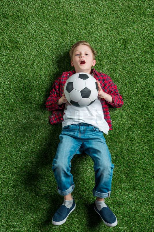 Top view of boy with soccer ball lying on green grass, stock photo