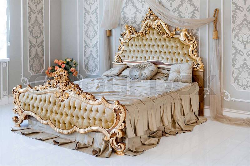 Luxury bedroom in light colors with golden furniture details. Big comfortable double royal bed in elegant classic interior, stock photo