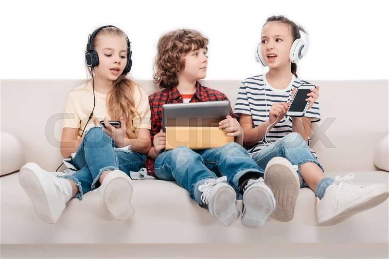 Cute kids using digital devices, listening music while sitting on couch, stock photo