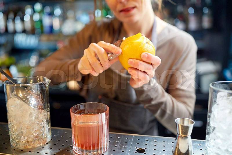Alcohol drinks, people and luxury concept - woman bartender with glass and peeler removing peel from orange and preparing cocktail at bar, stock photo