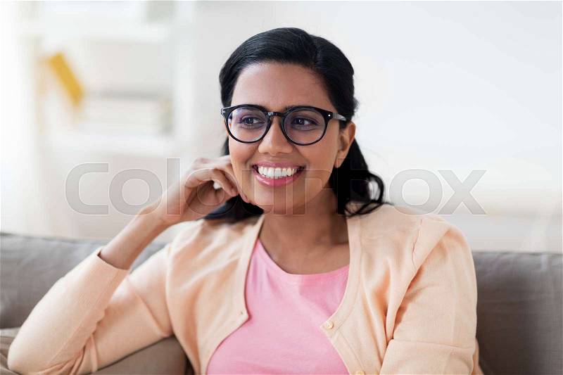 People, race, ethnicity and leisure concept - happy smiling young indian woman in glasses sitting on sofa at home, stock photo