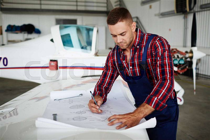 Handsome aircraft engineer reading blueprints while repairing plane in hangar, stock photo
