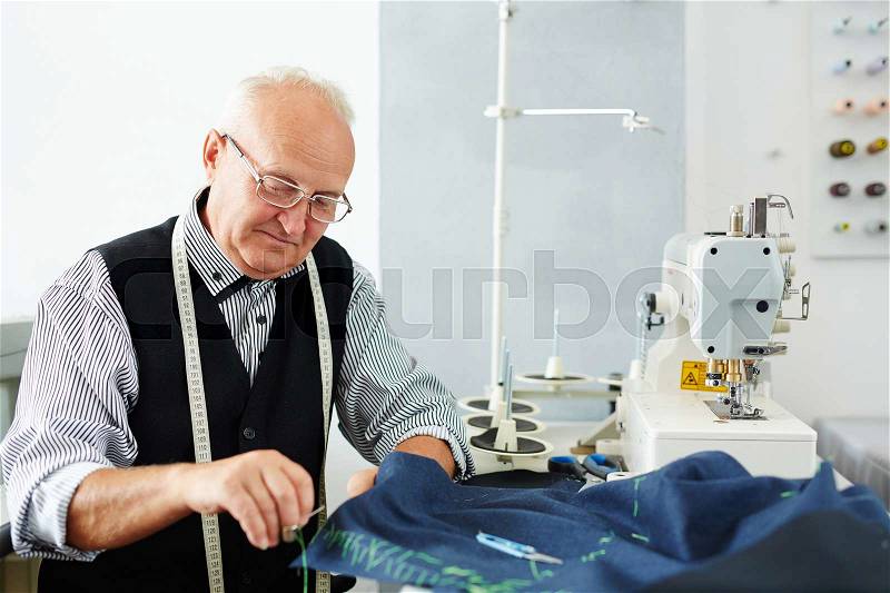 Portrait of old man working in tailoring studio making clothes at sewing machine and hand stitching cloth, stock photo