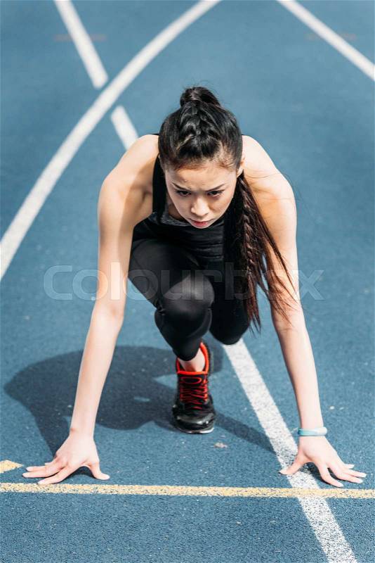 Concentrated young sportswoman in starting position on running track stadium, stock photo