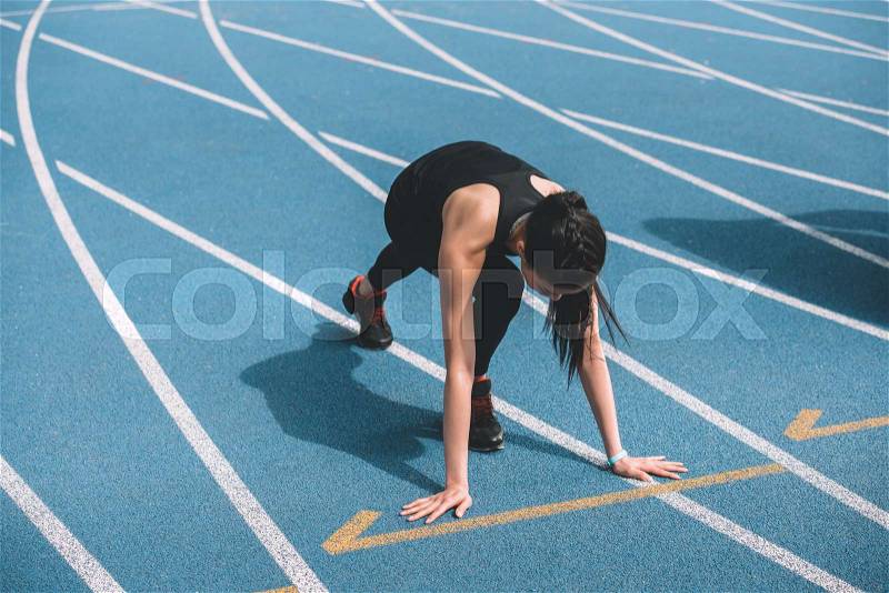 Concentrated young sportswoman in starting position on running track stadium, stock photo