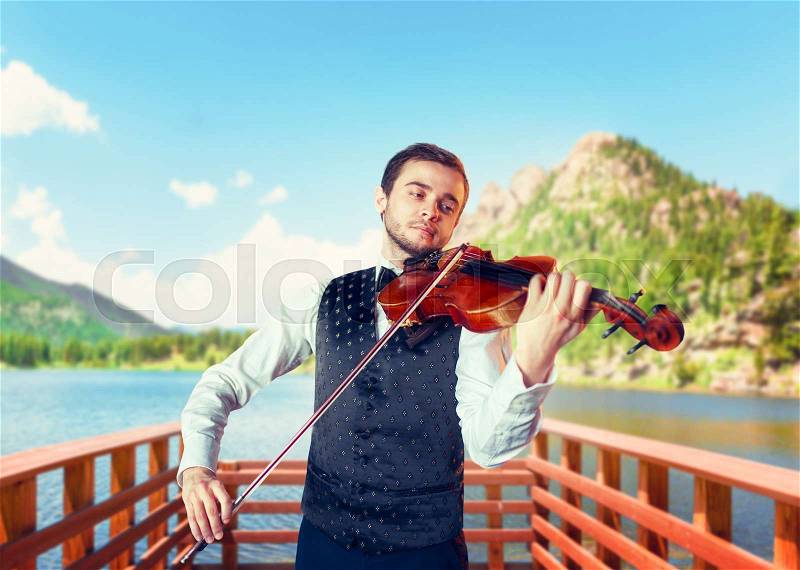 Male fiddler playing classical music on violin on wooden pier, lake and mountains on background. Violinist man with musical instrument plays on nature, stock photo