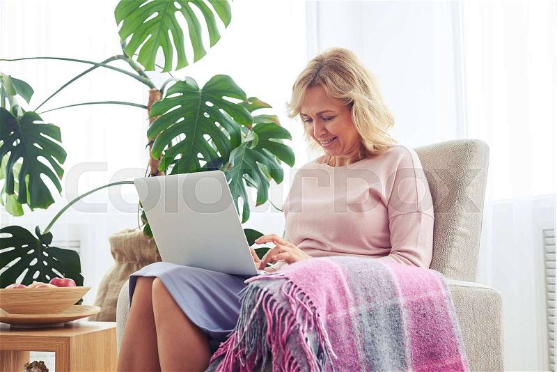 Mid shot of good-looking woman printing in laptop while sitting in armchair, stock photo