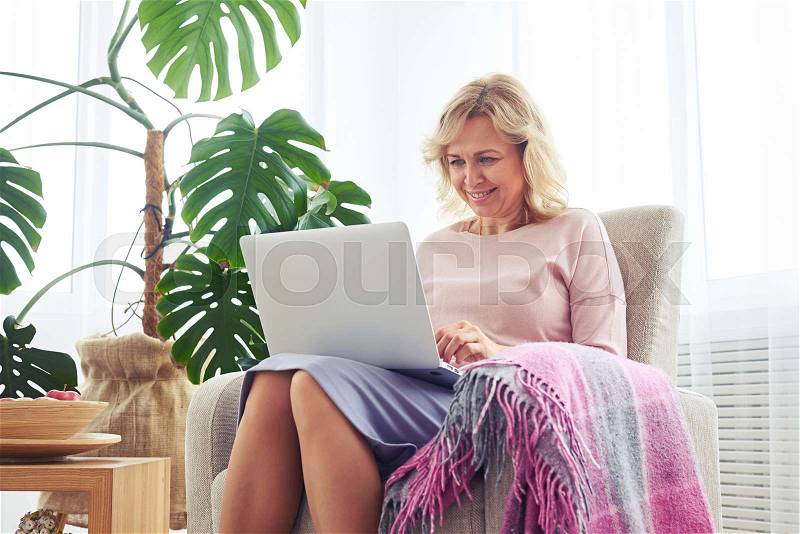 Mid shot of nice blonde printing in laptop while sitting in armchair, stock photo