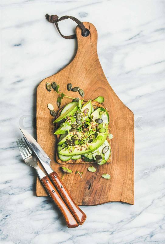 Healthy green veggie breakfast concept. Sandwich with fresh avocado, cucumber, kale, kress sprout, pumpkin seeds on rustic wooden board over marble background, top view. Vegetarian, detox food concept, stock photo