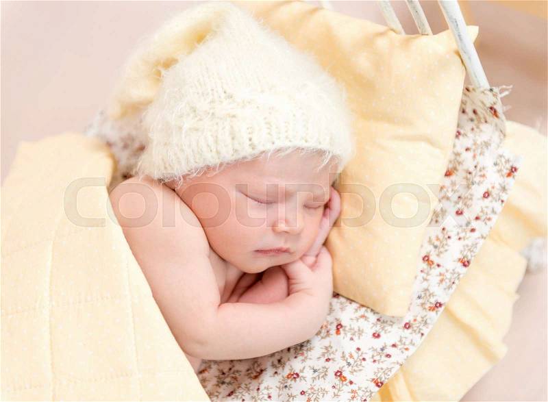 Lovely kid napping in cute yellow knitted hat and yellowish sheets in a bed, stock photo