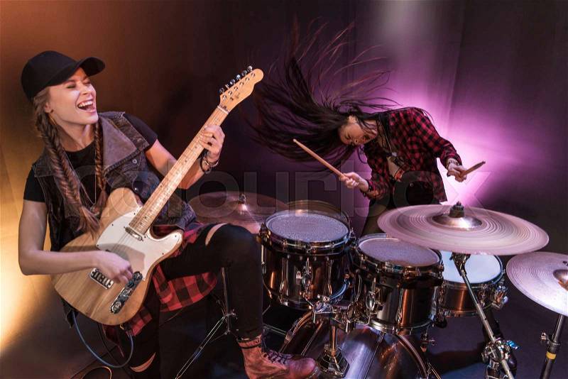 Excited young women with drums set and electric guitar performing rock concert on stage, stock photo